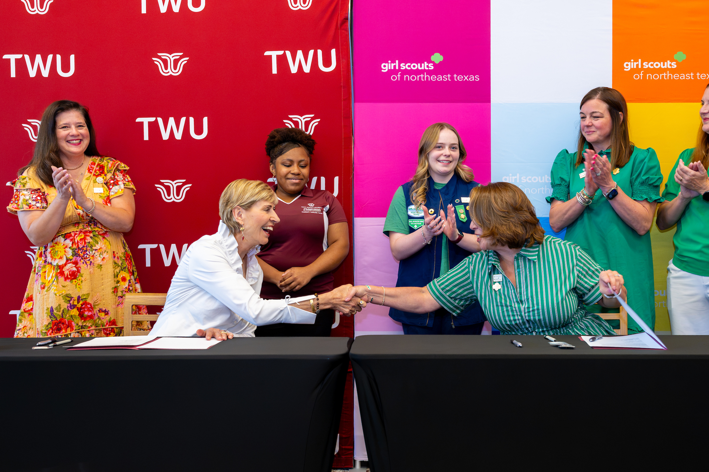 Two individuals shake hands at a table during a signing event, with supporters applauding behind them against branded backdrops.